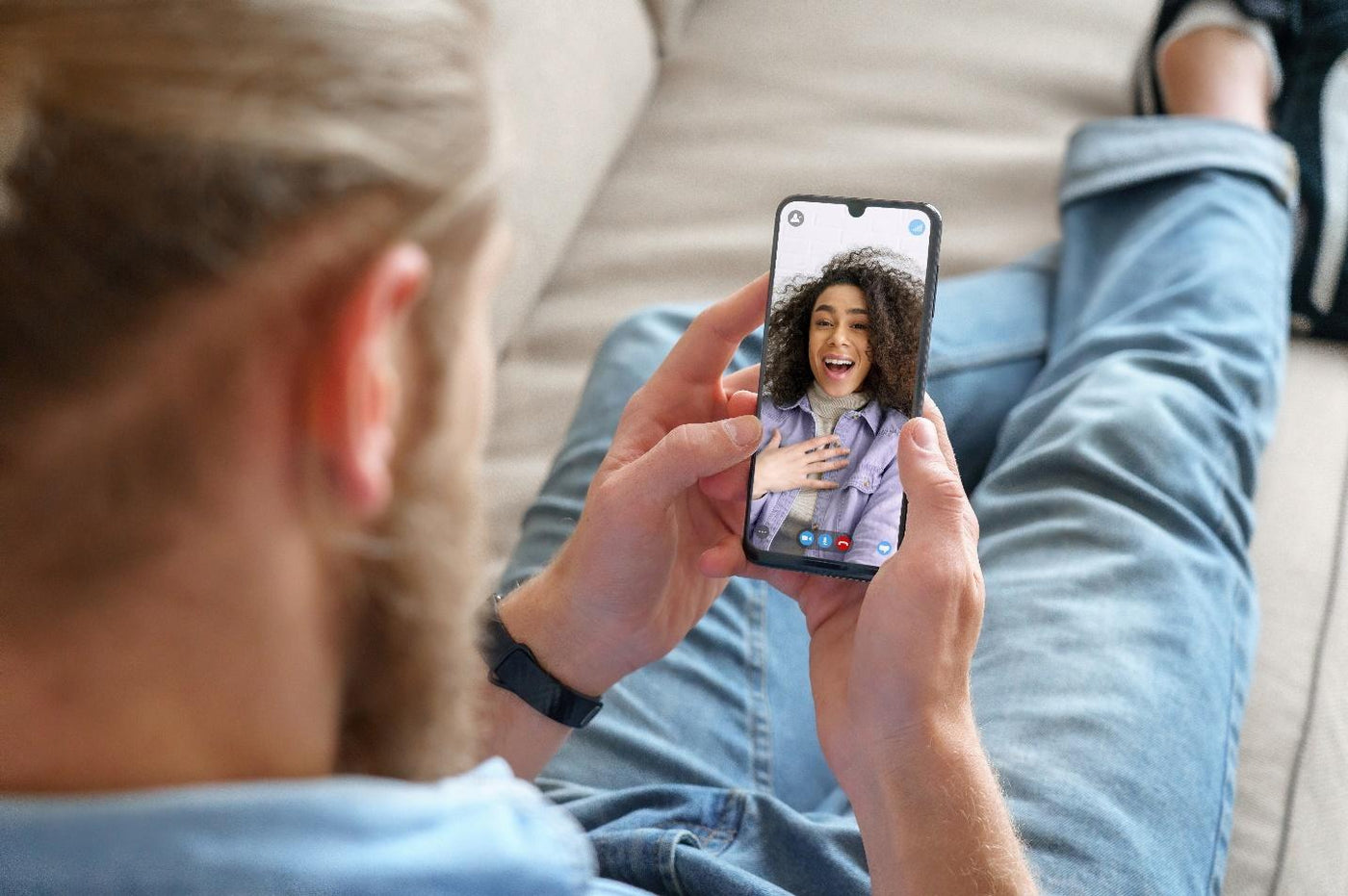 Young man sitting on sofa holding smartphone communicating with african girl friend