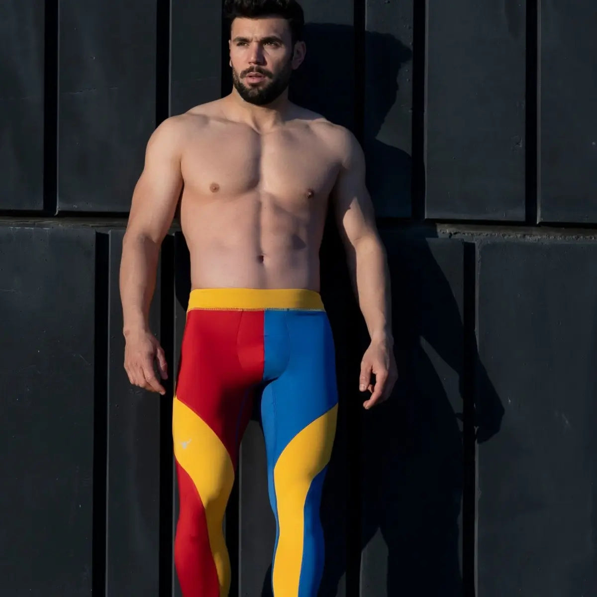 Red, blue, and yellow men's leggings worn by a shirtless man standing against a dark background.