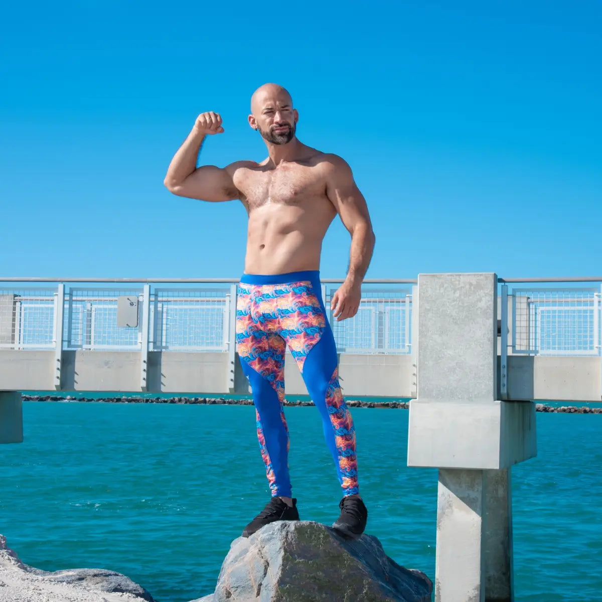Men's compression leggings showcased on a muscular man flexing in vibrant blue and orange leggings by the ocean