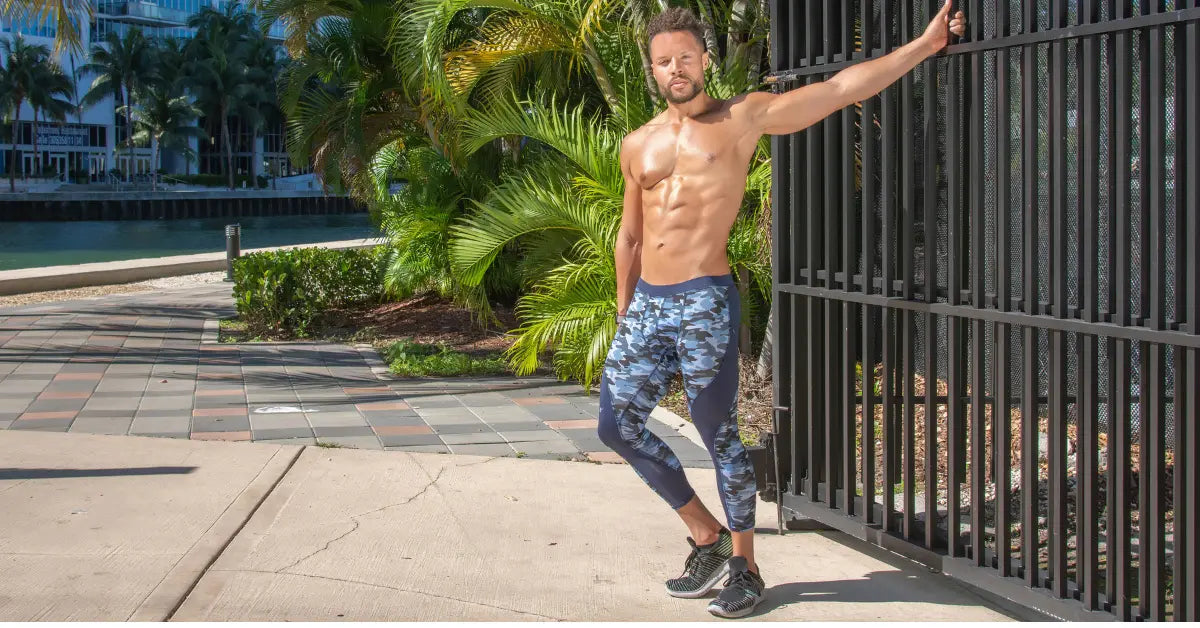 Printed workout leggings on a shirtless man standing outdoors in front of palm trees and a black metal gate.